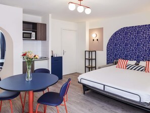 Habitación de hotel compacta con cama, una mesa redonda con sillas, kitchenette y una pared decorativa.