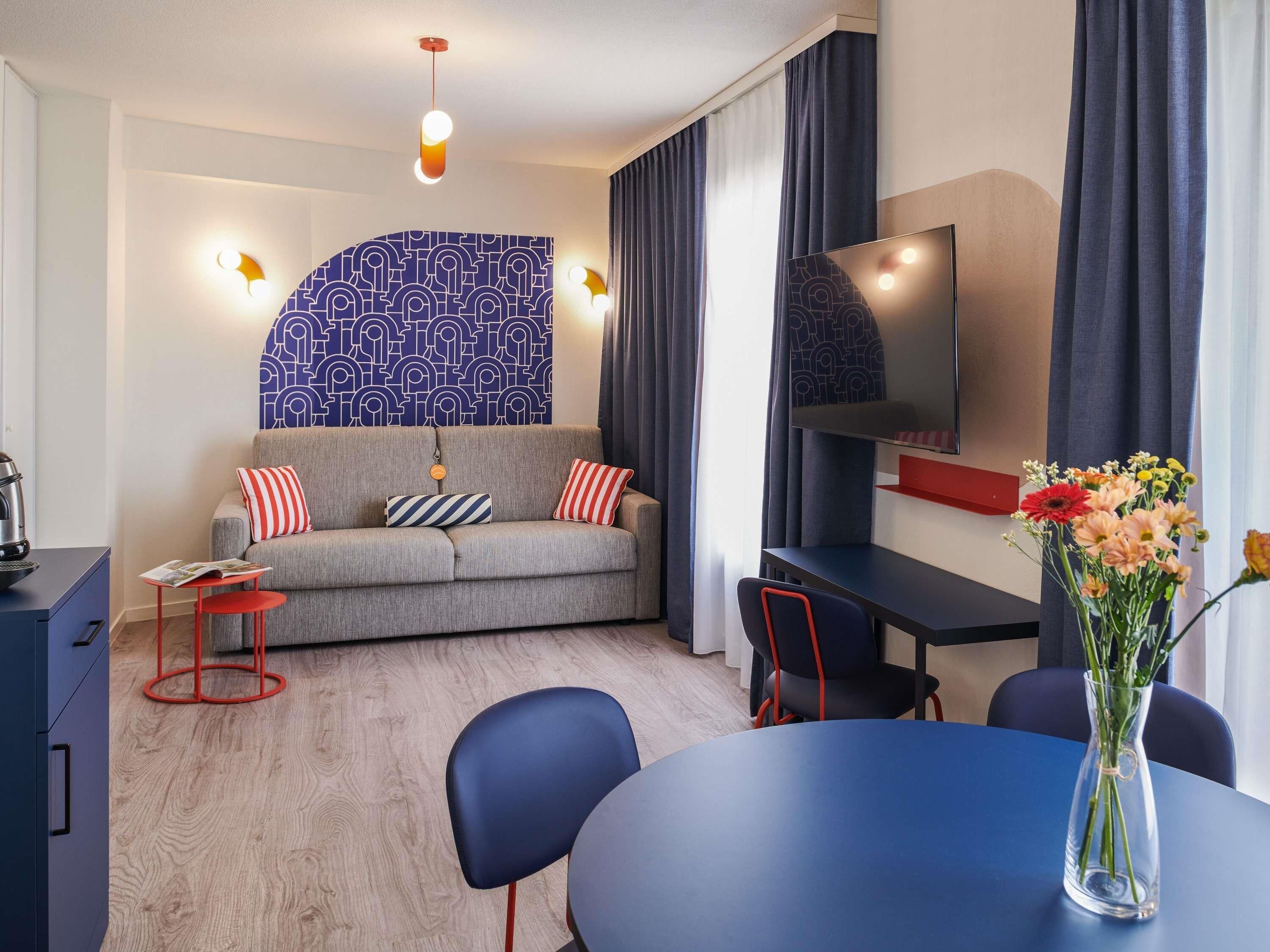 Ein modernes Hotelzimmer mit einer Couch, einem Esstisch, einem Fernseher und einer Vase mit Blumen.