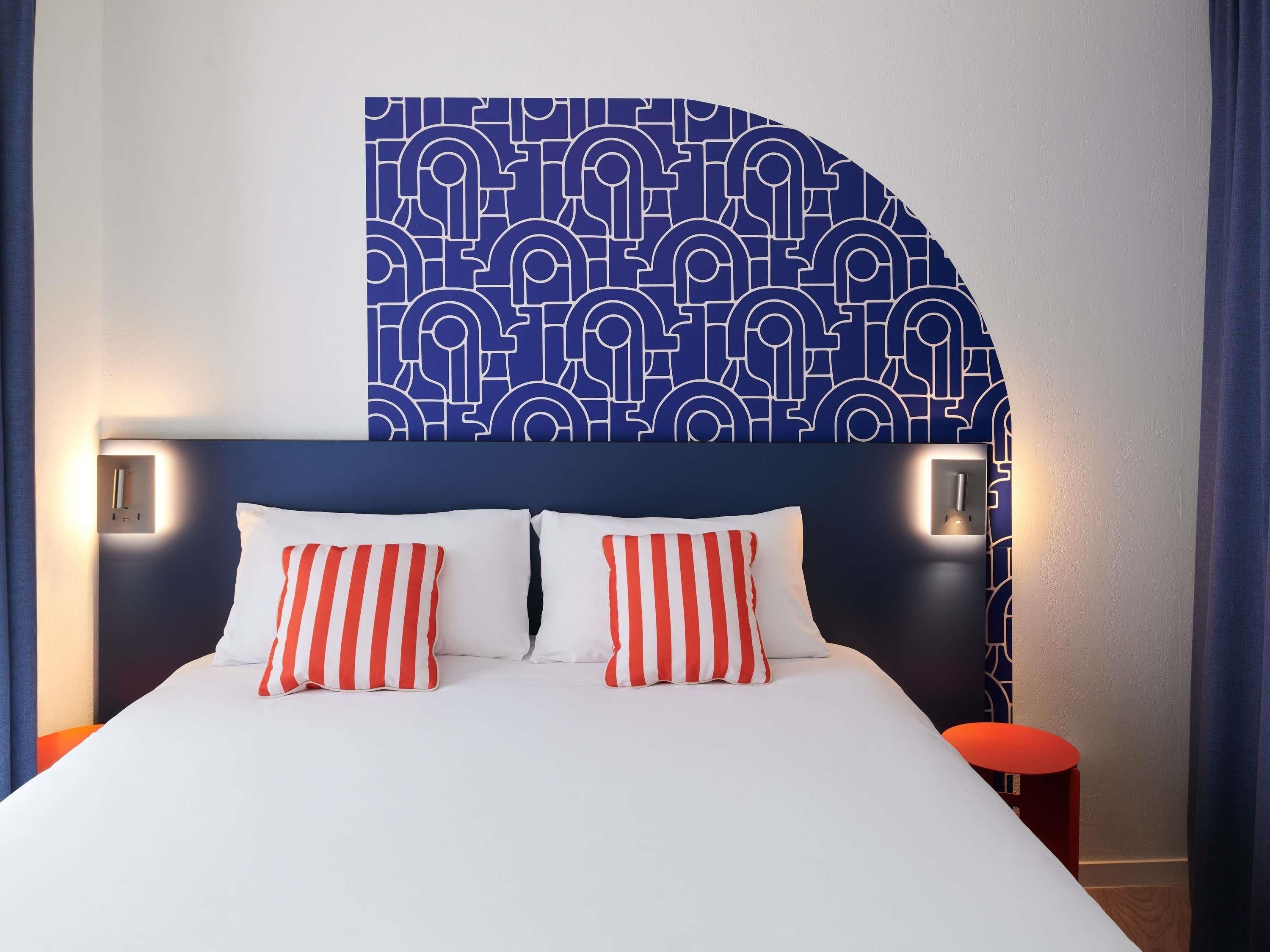 A hotel room with a bed, two pillows, a headboard, bedside lamps, and a wall with a geometric pattern.