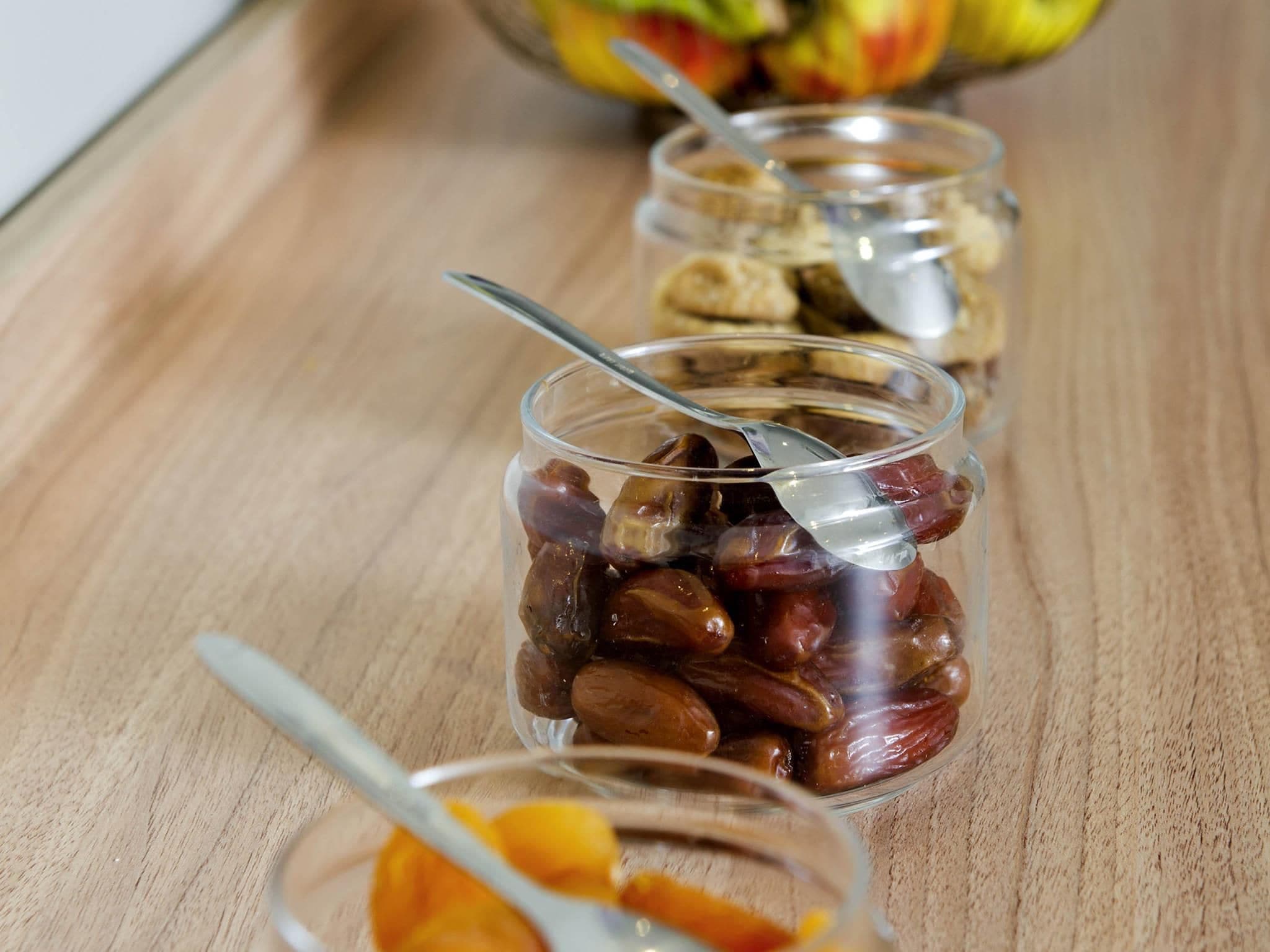 Three glass jars with a spoon, containing what appears to be dried fruits, on a wooden surface.