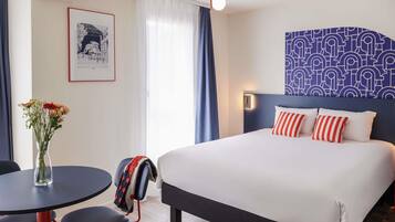 Habitación de hotel con cama, escritorio, silla, un jarrón con flores y un diseño decorativo en la pared.
