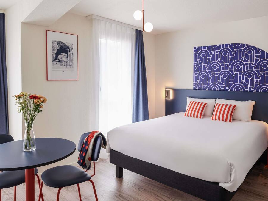 Quarto de hotel com cama, escrivaninha, cadeira, um vaso de flores e um papel de parede com padrão.