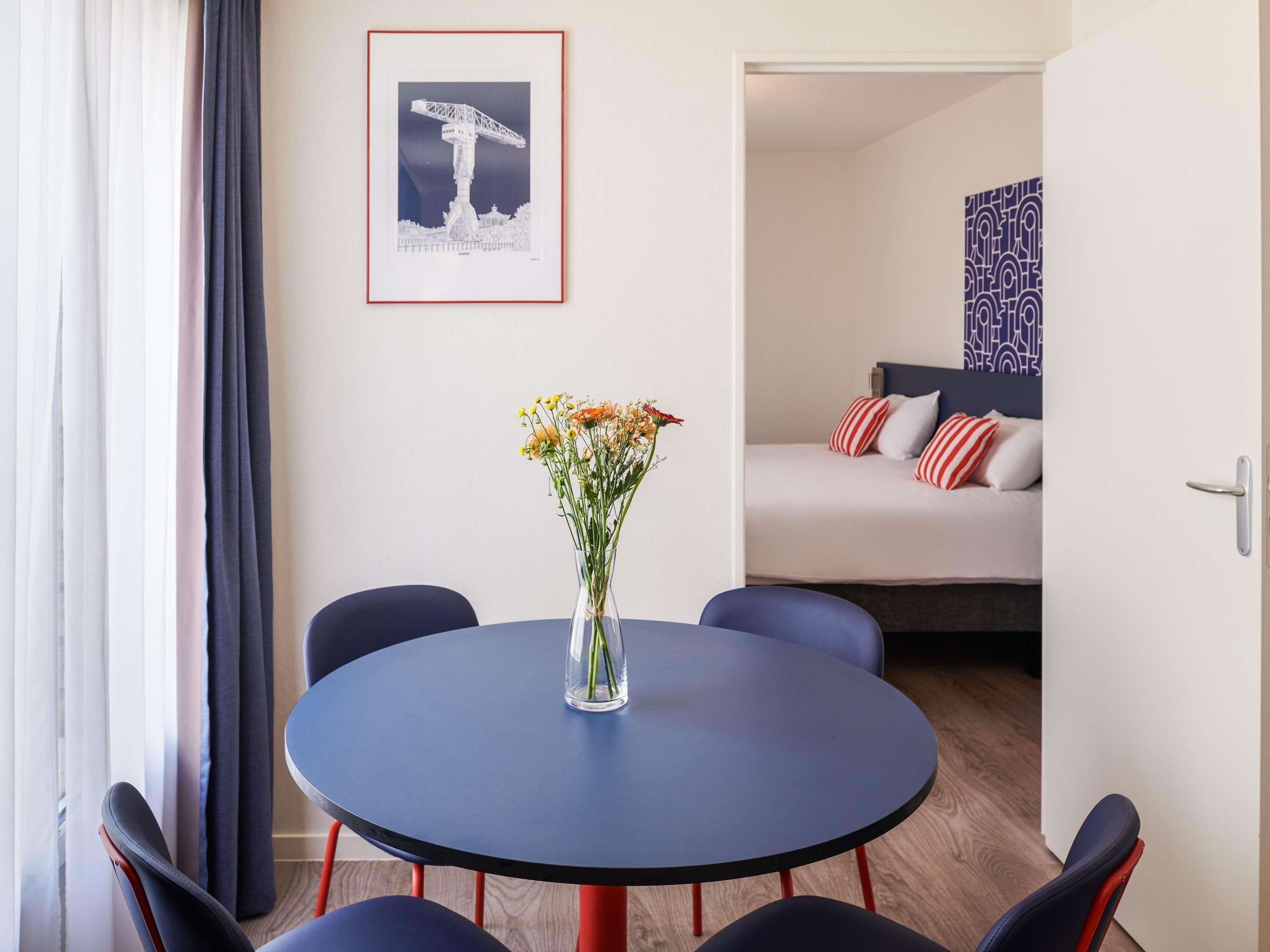 Een kamer met een ronde tafel, stoelen, een vaas met bloemen, een bed met kussens en een ingelijst schilderij aan de muur.