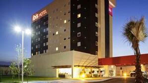 Um hotel de vários andares com fachada plana, um destaque para a placa 'ibis HOTEL' e uma área de entrada bem iluminada.