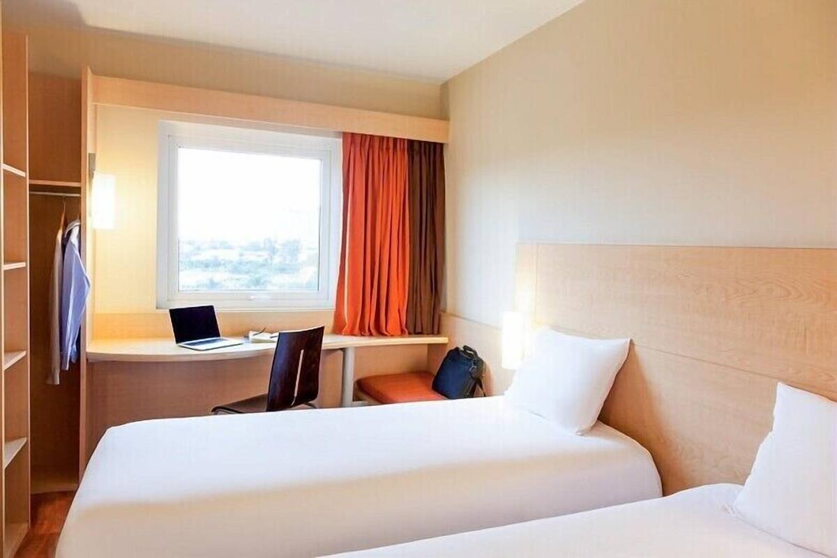 Ein Hotelzimmer mit zwei Betten, einem Schreibtisch, einem Laptop, einem Stuhl und einem Fenster mit Vorhängen.