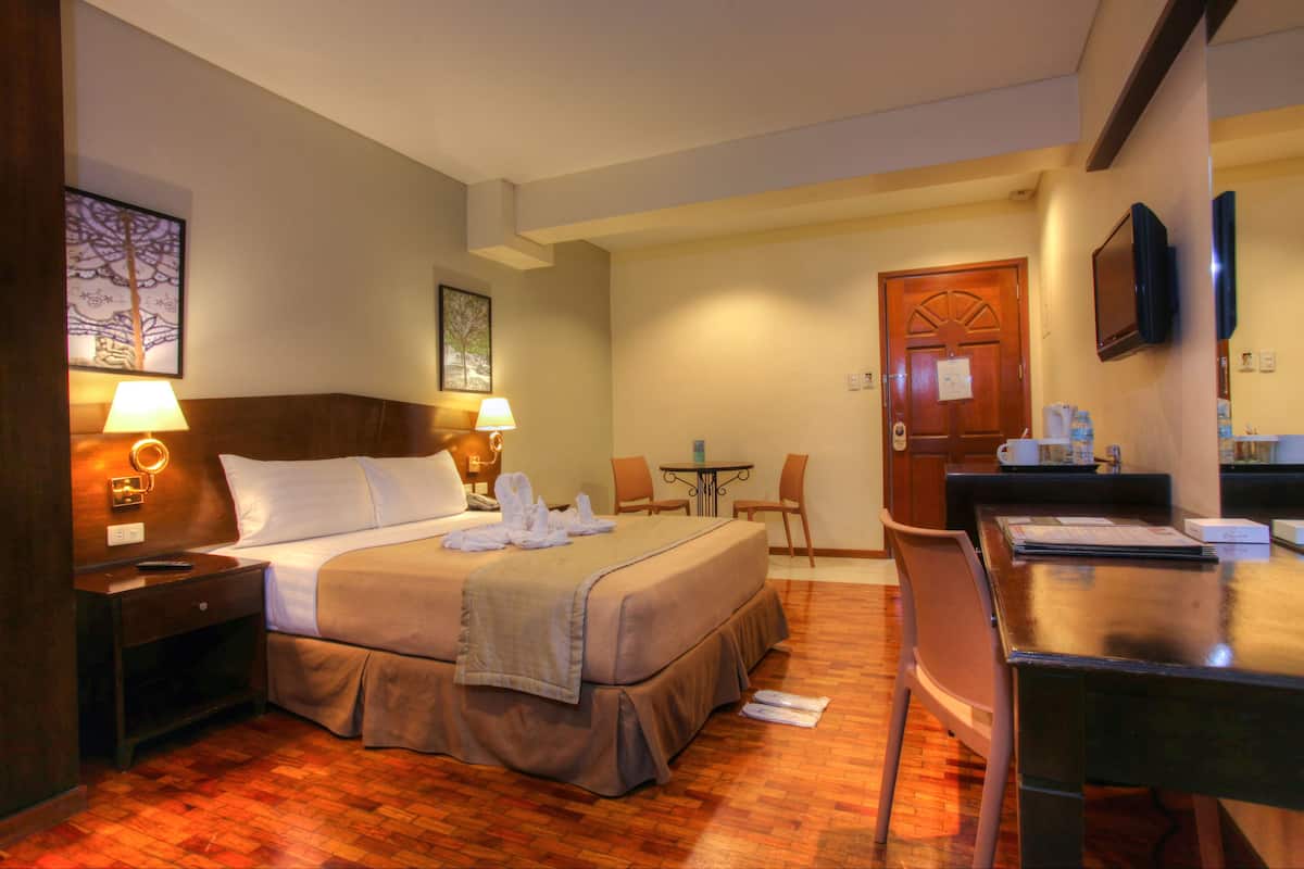 A hotel room with a bed, bedside lamps, a desk, a chair, a small table, and a TV mounted on the wall.