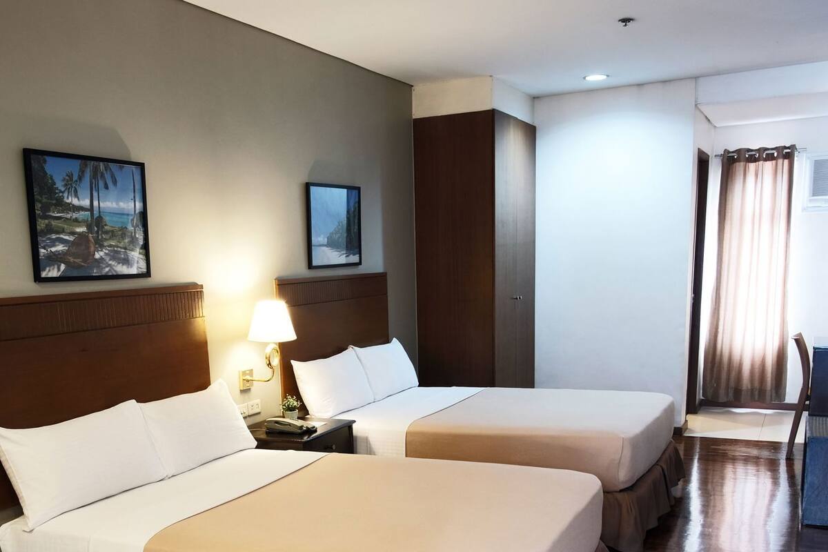 A hotel room with two beds, a nightstand, a lamp, a painting, and a view of the outdoors.