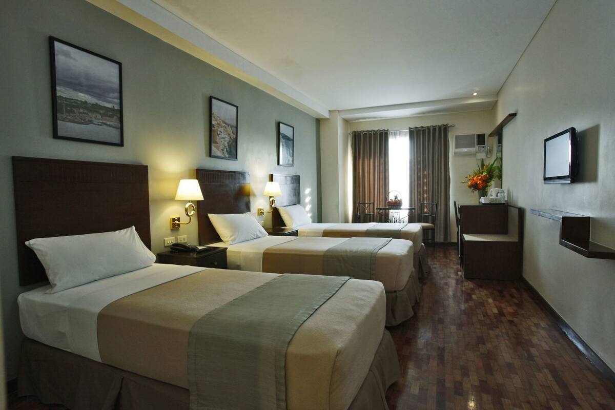 A hotel room with two beds, a small table, a TV, and a bathroom visible in the background.