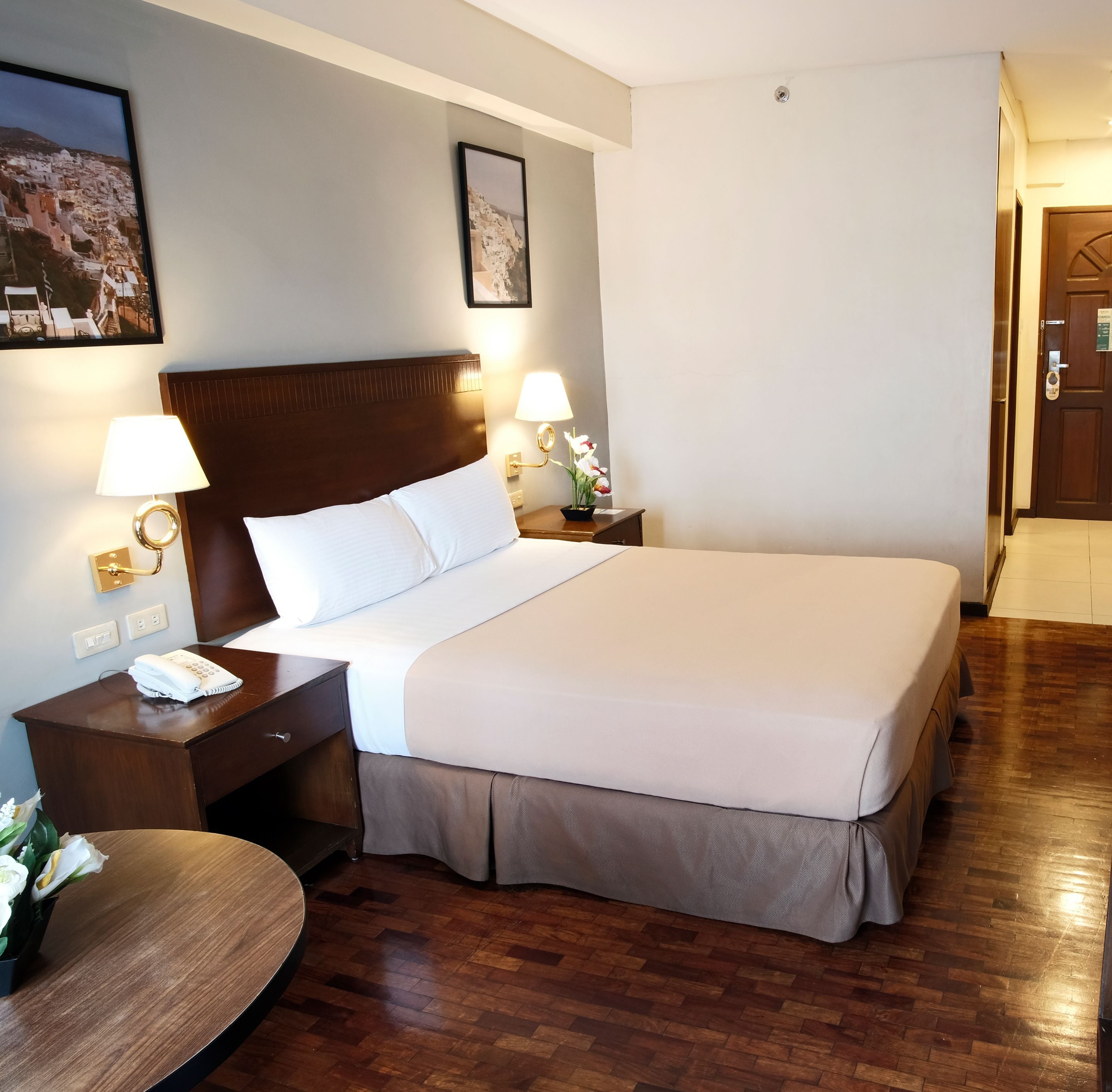 A hotel room with a large bed, wooden headboard, two bedside tables with lamps, a telephone, and a small table with a flower arrangement.