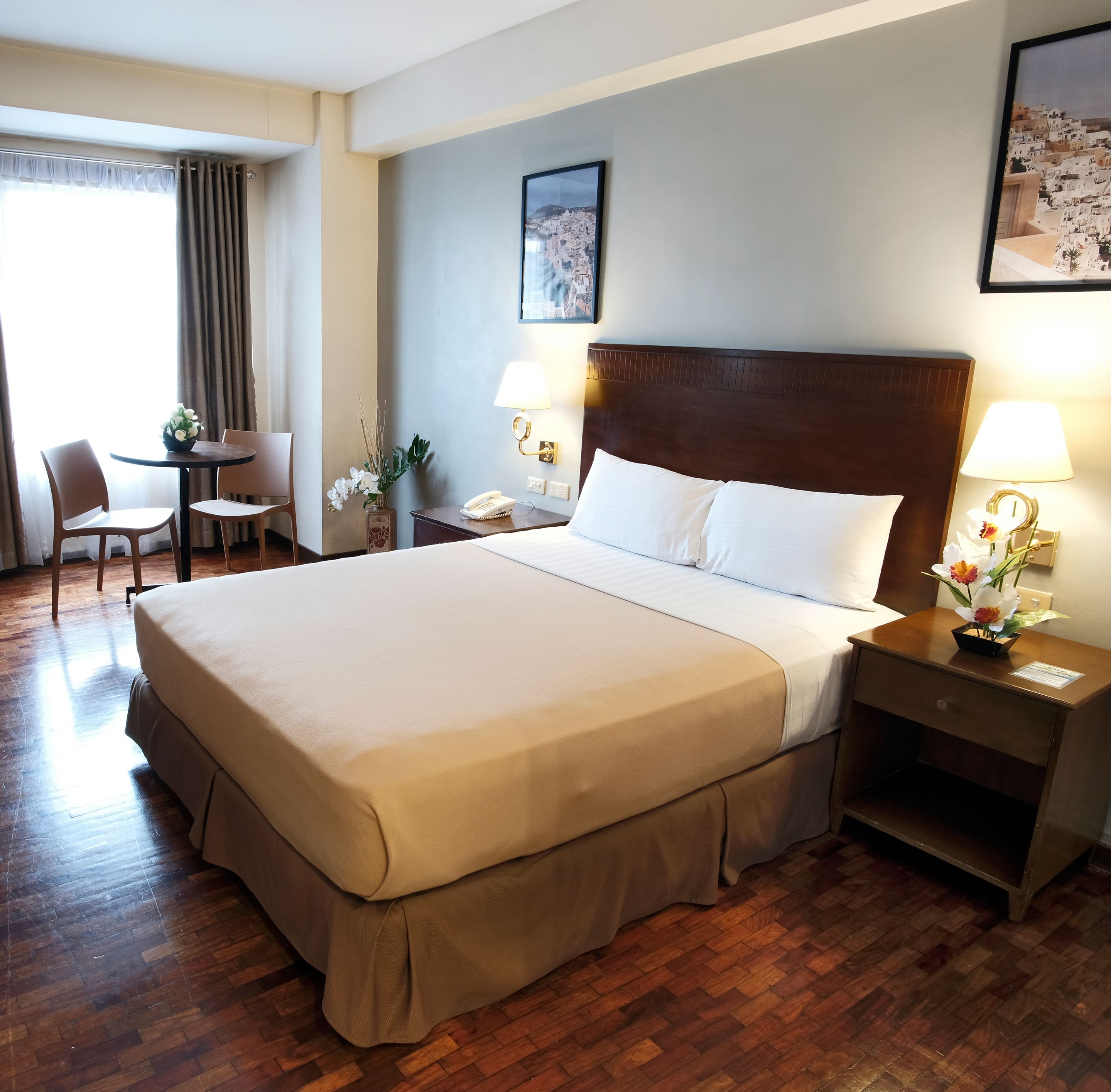 A hotel room with a large bed, a desk with a chair, a small table with a vase of flowers, and a wall-mounted lamp.