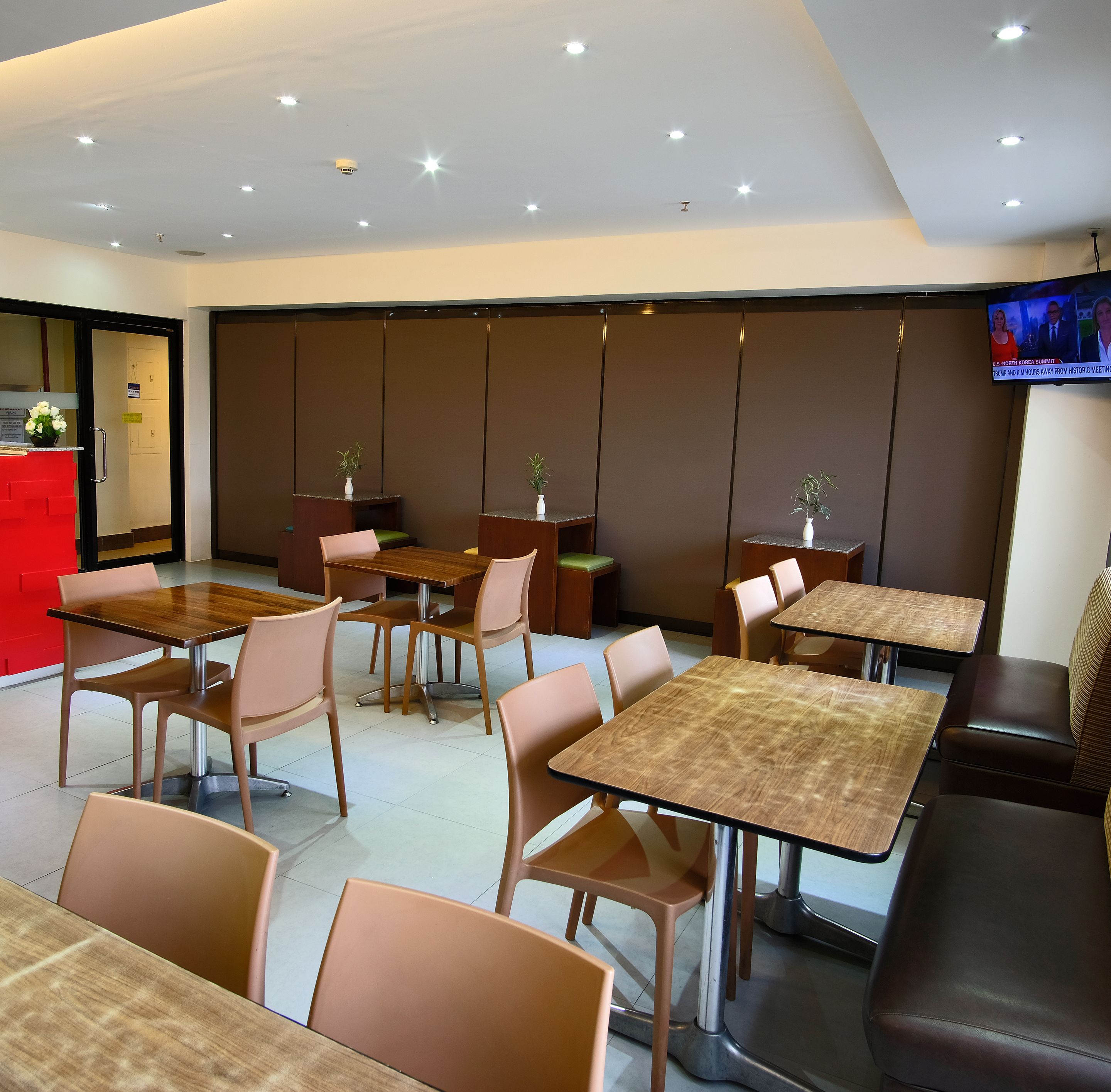 A modern dining area with multiple tables and chairs, a reception desk, and a television screen displaying news.
