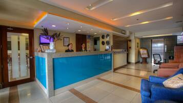 A hotel reception area with a blue reception desk, two receptionists, a clock, and a seating area with blue sofas.