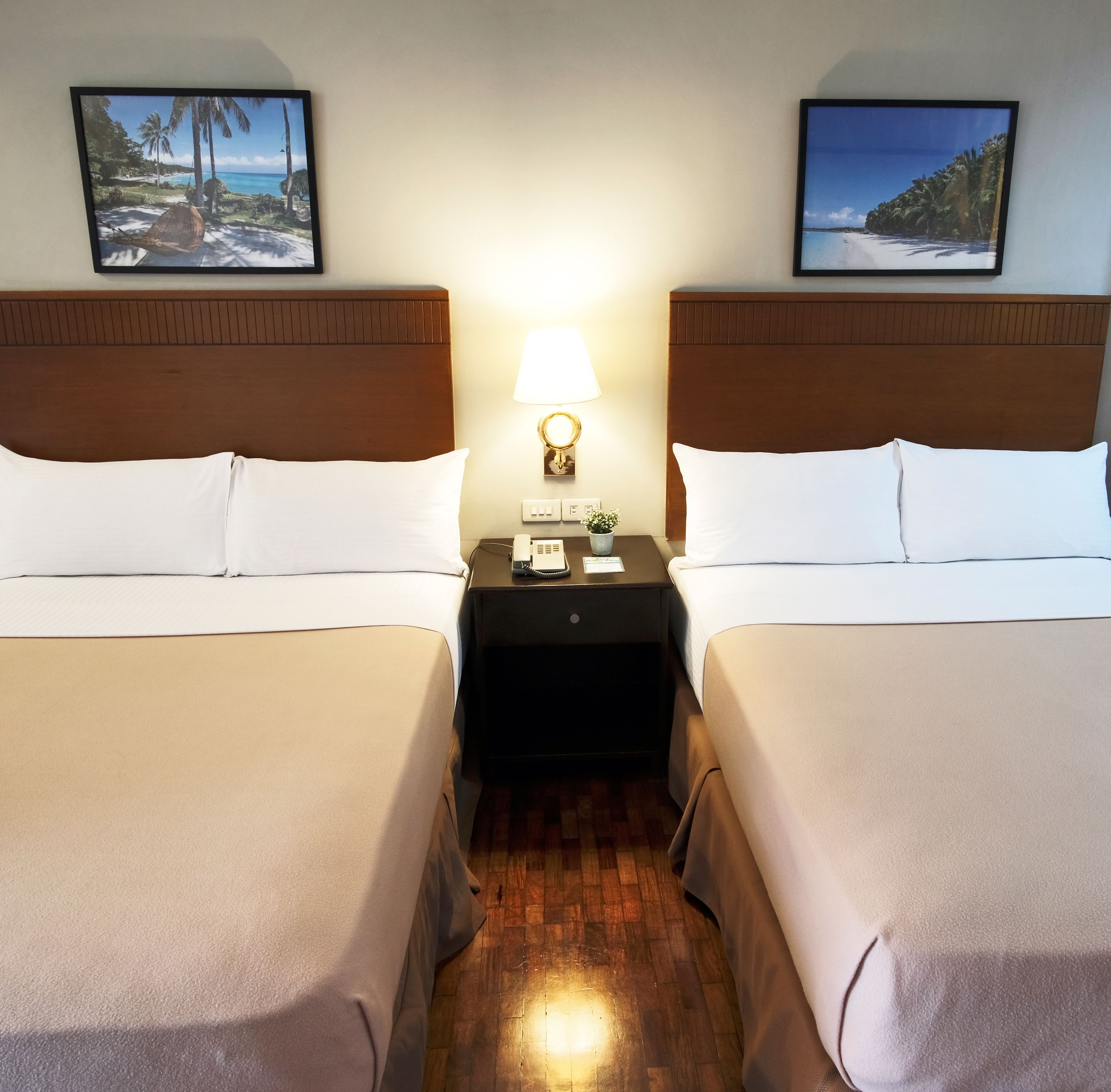 A hotel room with two beds, a nightstand with a lamp, and two framed pictures on the wall.