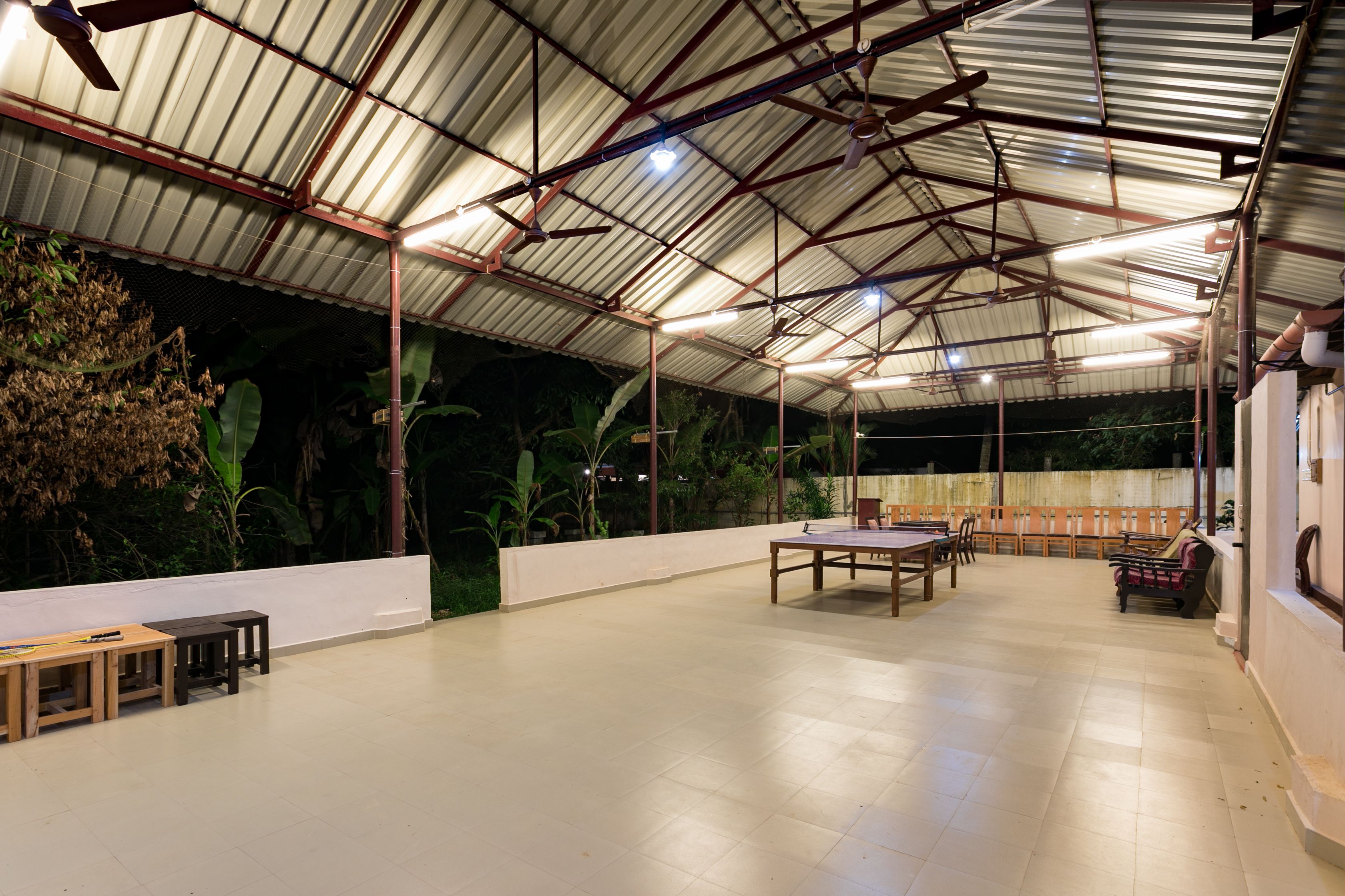 An indoor space with a high ceiling, metal beams, and a tiled floor. There are tables and chairs arranged for dining or socializing.