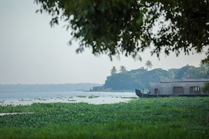 A traditional boat house by a calm water body with lush greenery and distant palm trees. - ILLIKKALAM Lakeside Cottages Kumarakom Kerala (Kottayam)