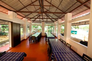 A covered dining area with tables and chairs, large windows, and a view of greenery outside. - ILLIKKALAM Lakeside Cottages Kumarakom Kerala (Kottayam)