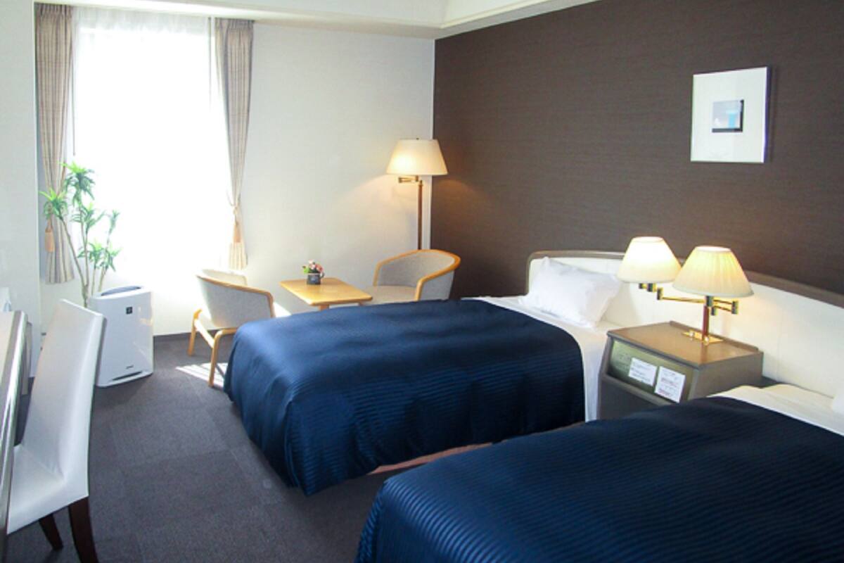 A hotel room with two beds, a desk, a chair, a lamp, and a plant.