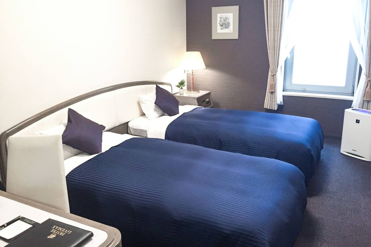 A hotel room with two beds, a desk, a lamp, a small table with a plant, and a window with curtains.