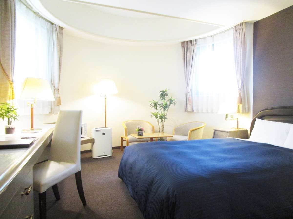 Habitación de hotel con cama, escritorio, sillas, una mesita, lámpara y planta en maceta.
