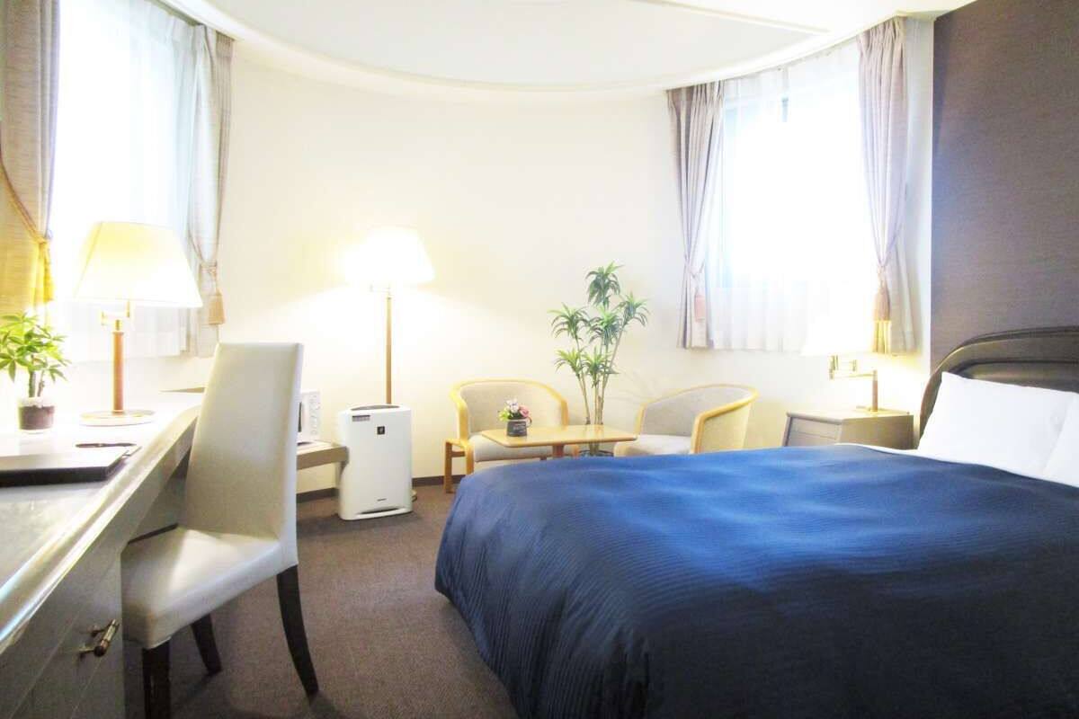 Une chambre d’hôtel avec un lit, un bureau, des chaises, une petite table, une lampe et une plante en pot.