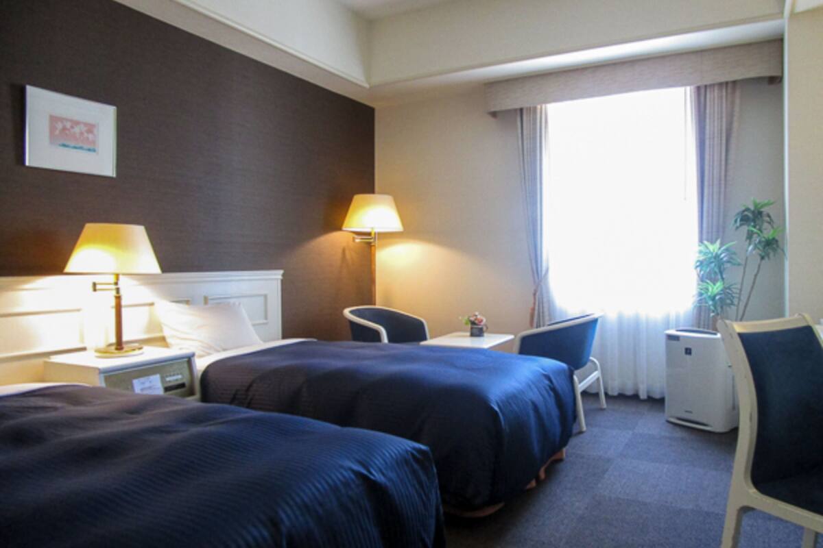 A hotel room with two beds, a desk, a chair, a lamp, a plant, and a window with curtains.