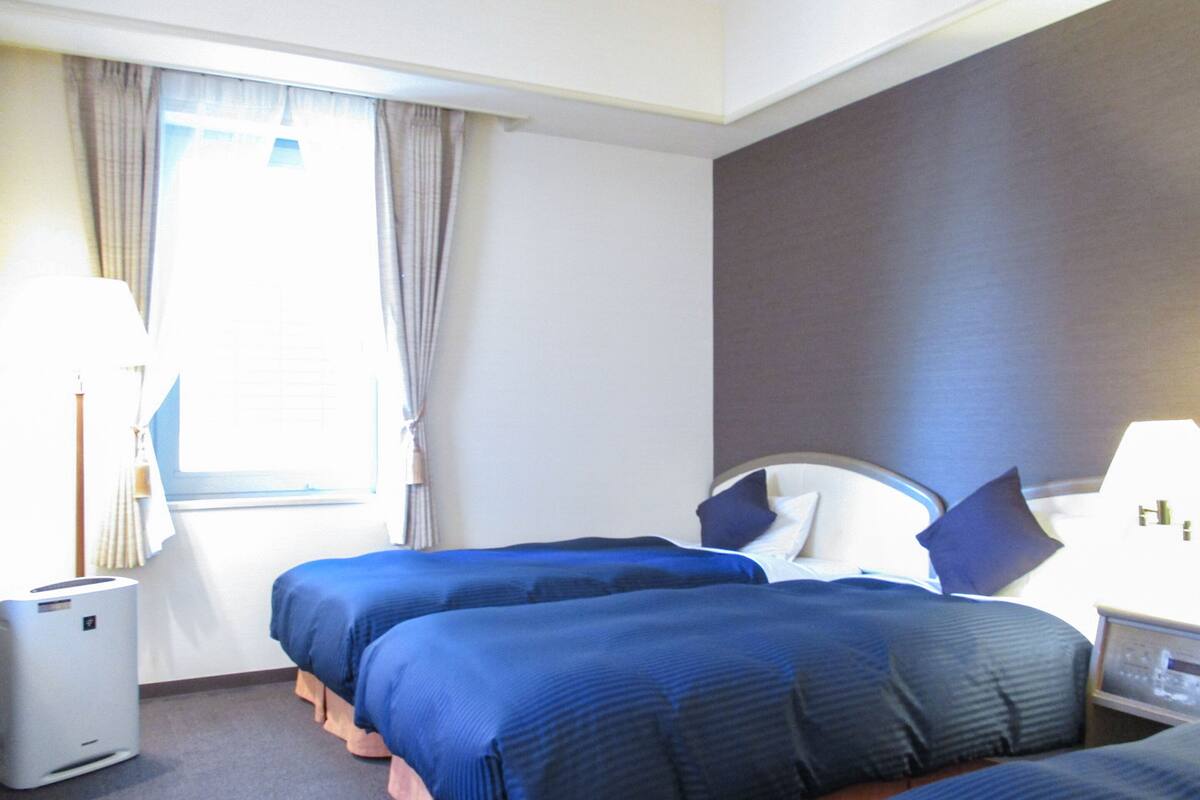 A hotel room with two beds, a bedside table, a lamp, and a window with curtains.