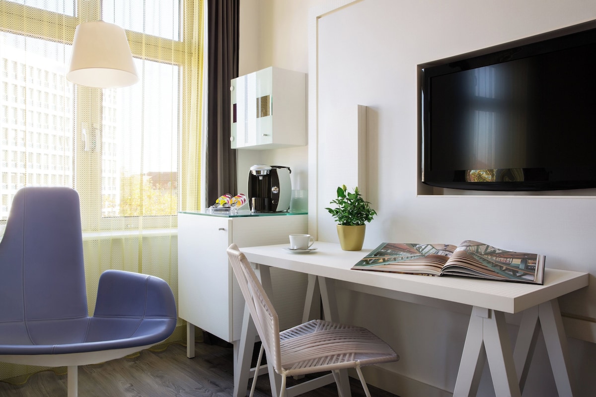 Une chambre d’hôtel moderne avec un bureau blanc, une chaise bleue, une autre chaise blanche, une machine à café, une plante en pot et un téléviseur à écran plat.