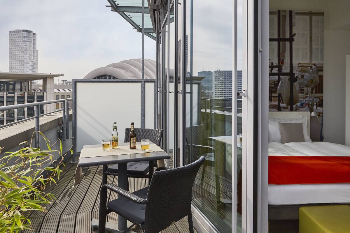 Un balcon avec une table et des chaises, un lit avec une couverture rouge et une vue sur la ville.