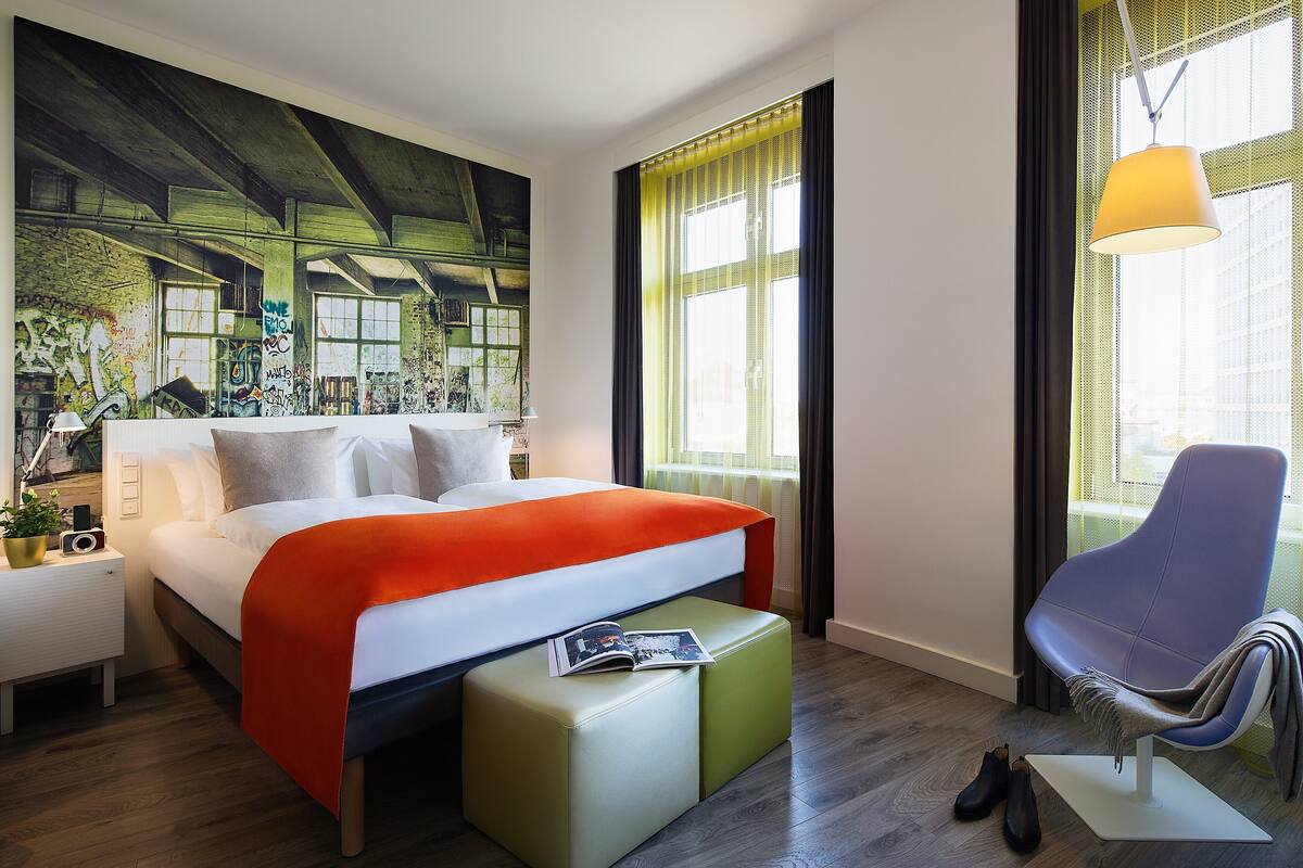 Une chambre d’hôtel moderne avec un grand lit, un fauteuil de lecture, une petite table et une grande fenêtre avec des rideaux.