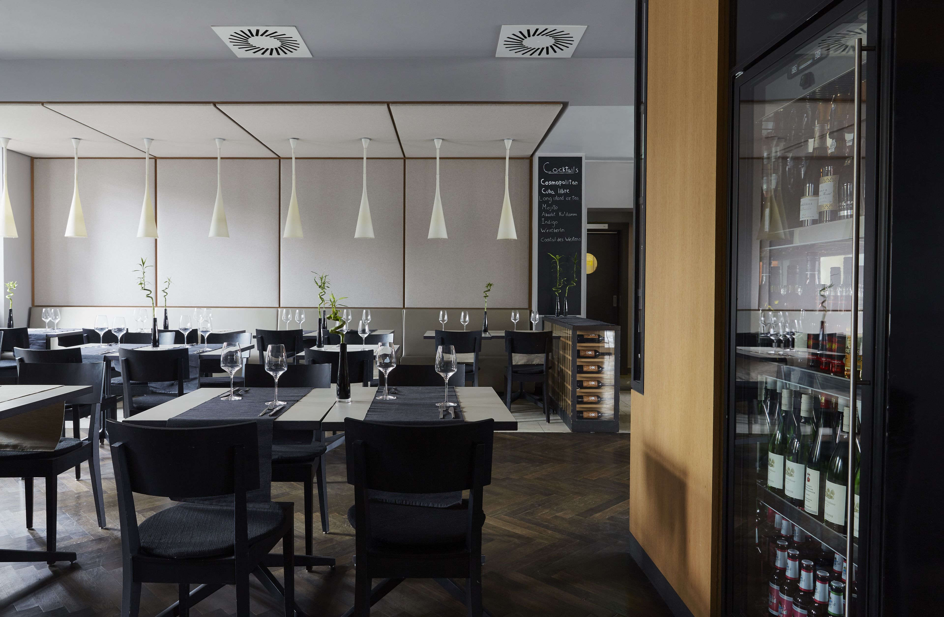 A modern restaurant interior with multiple dining tables, black chairs, hanging pendant lights, and a wine refrigerator stocked with bottles.