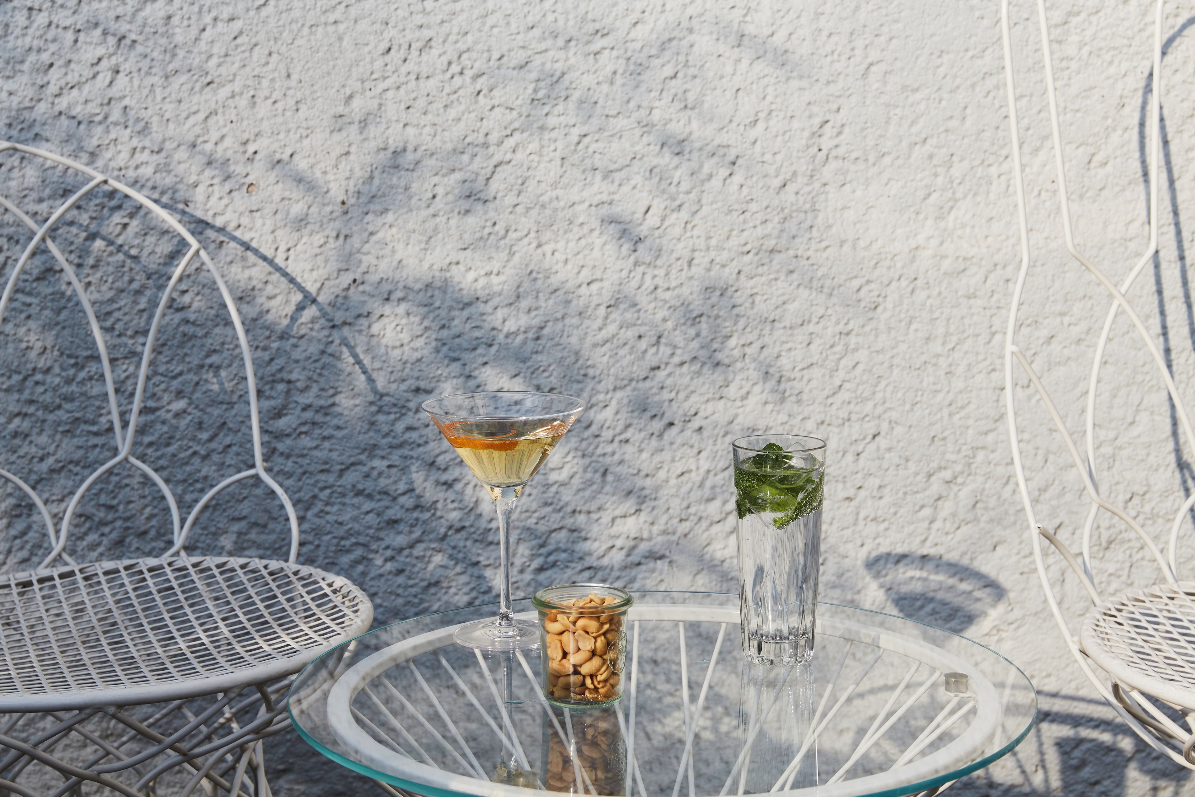 Mesa de vidro com duas cadeiras, uma taça de martini, um copo alto com bebida e um pote de nozes.