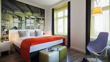 Una habitación de hotel moderna con una cama grande, un sillón de lectura, una mesita y un ventanal con cortinas.