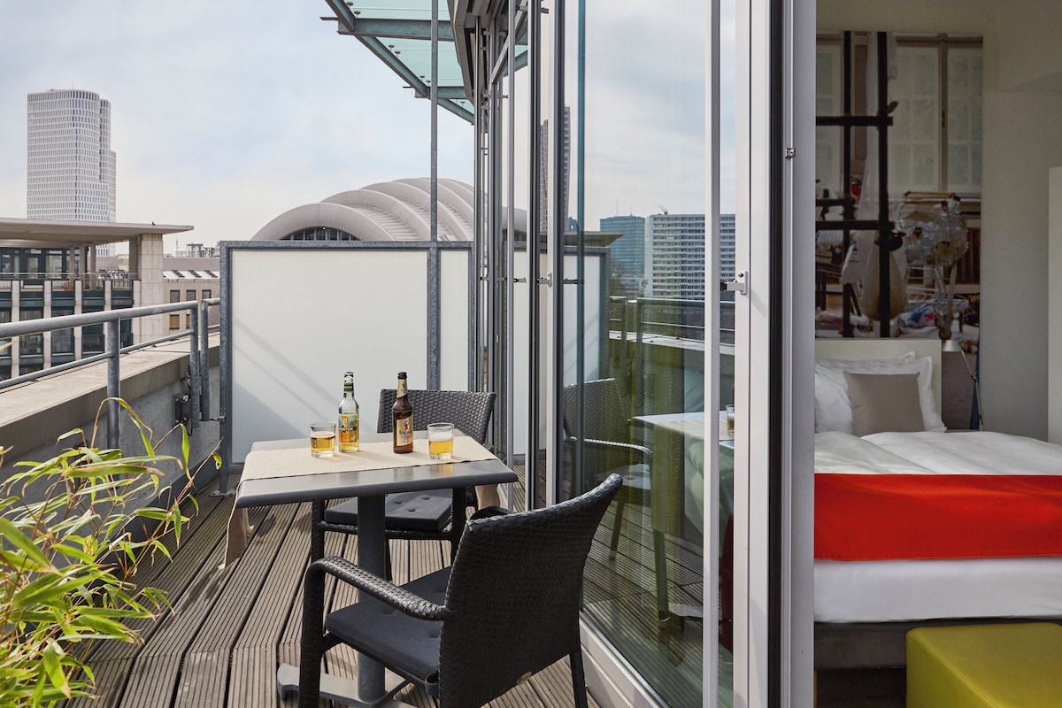 Un balcon avec une table et des chaises, un lit avec une couverture rouge et une vue sur la ville.