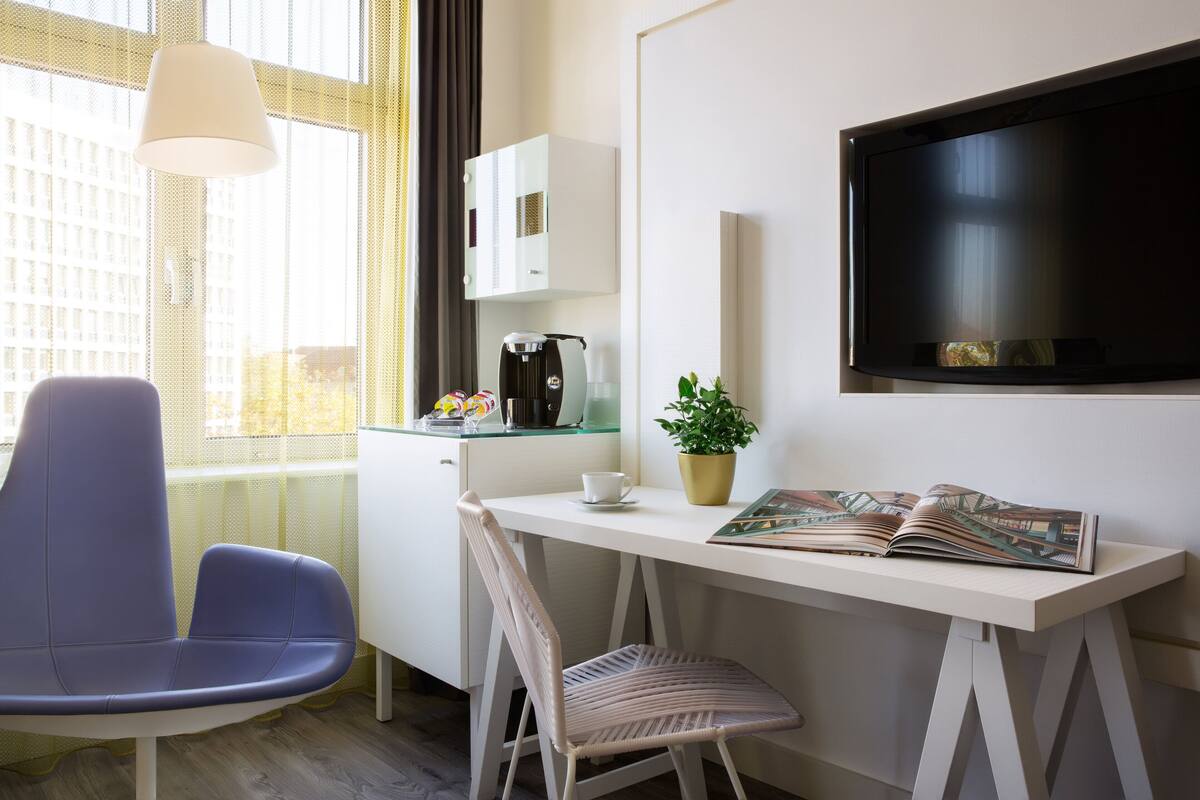 Une chambre d’hôtel moderne avec un bureau blanc, une chaise bleue, une autre chaise blanche, une machine à café, une plante en pot et un téléviseur à écran plat.