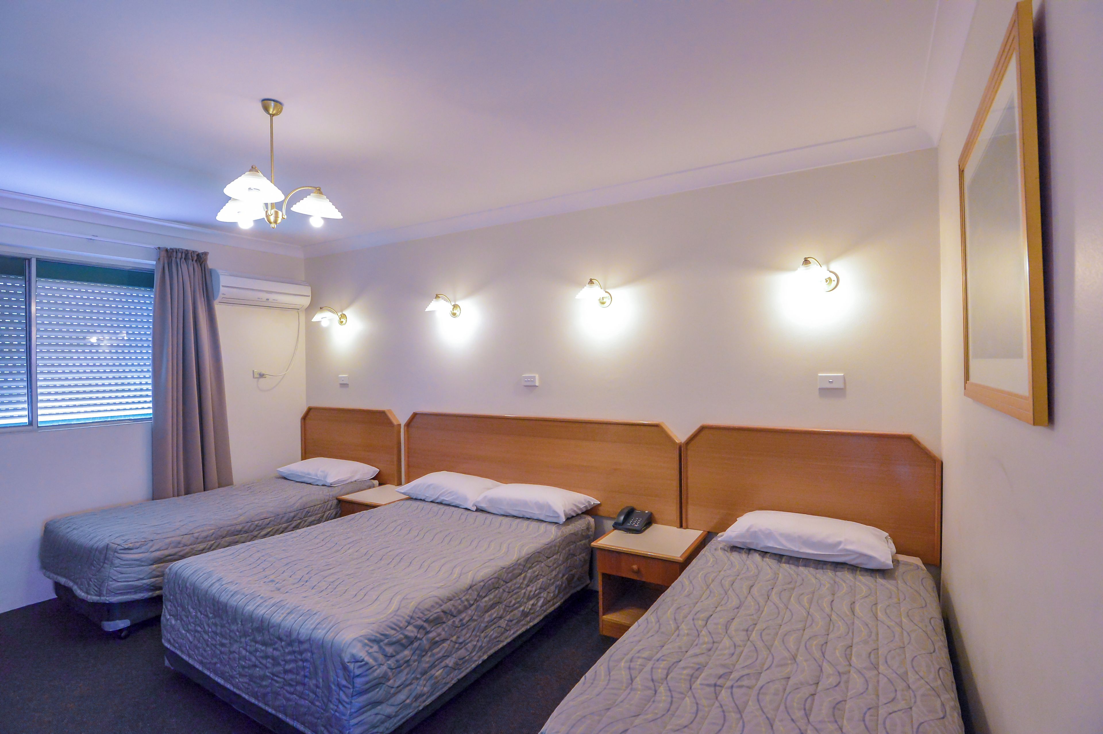 A hotel room with three single beds, a bedside table with a phone, and a window with blinds.