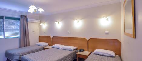 A hotel room with three single beds, a bedside table with a phone, and a window with blinds.