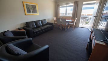 A living room with a black leather sofa, a dining table with chairs, a television, and a balcony with a view.
