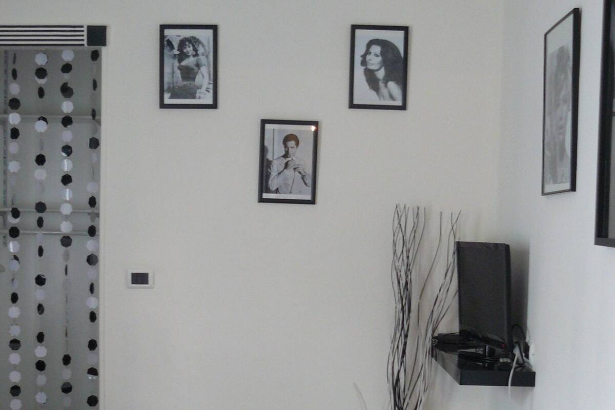 A hotel room with a bed, a desk, a chair, a TV, and three framed pictures on the wall.