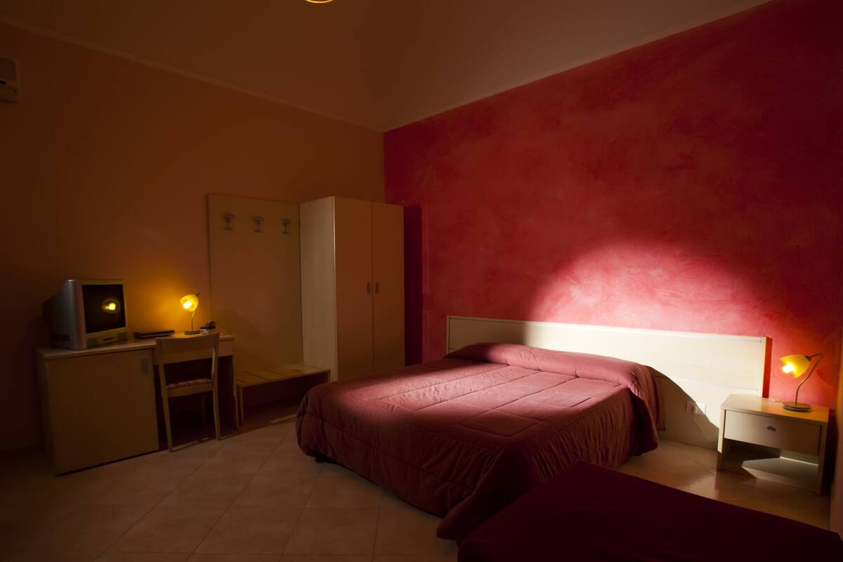 A bedroom with a bed, a desk with a computer, a wardrobe, and a nightstand with a lamp.
