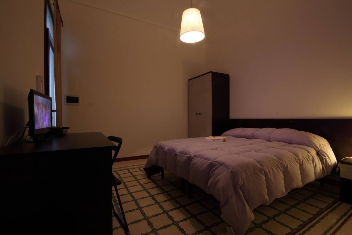 A hotel room with a bed, a desk, a chair, a wardrobe, and a hanging lamp.