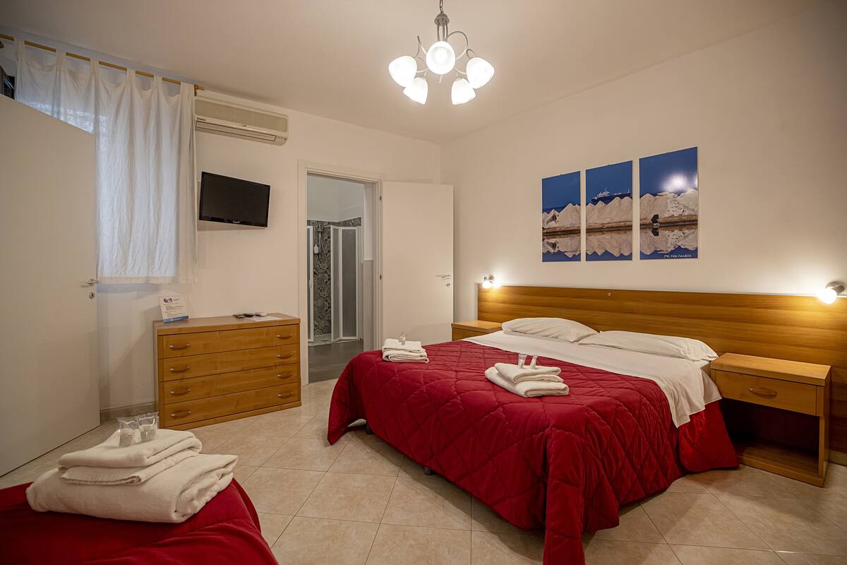 A bedroom with a bed, a wooden nightstand, a dresser, a TV, and a door leading to a bathroom.