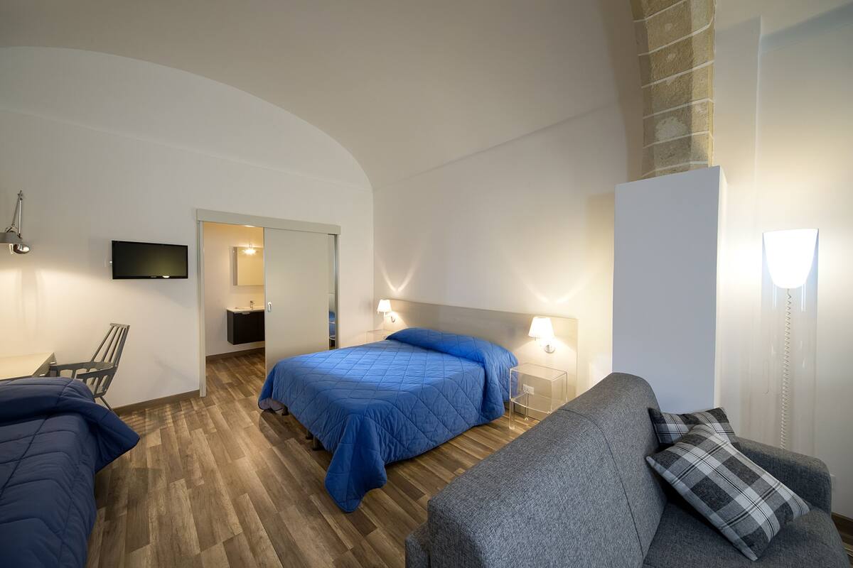 A modern hotel room with a bed, a sofa, a desk, and a TV.