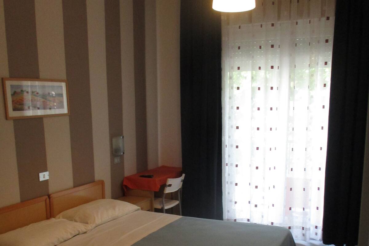 Una habitación de hotel con una cama grande, un escritorio, una silla, un cuadro en la pared y una ventana con cortinas.