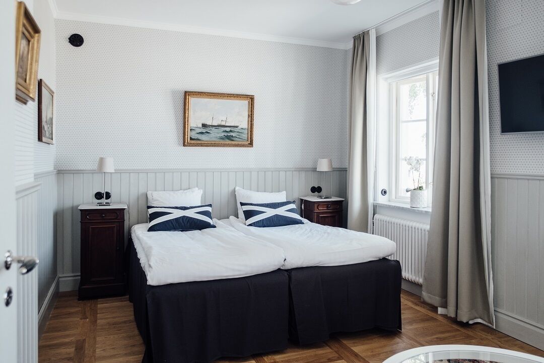A double bed with white and navy bedding, two bedside tables with lamps, a framed picture on the wall, a window with curtains, and a radiator.