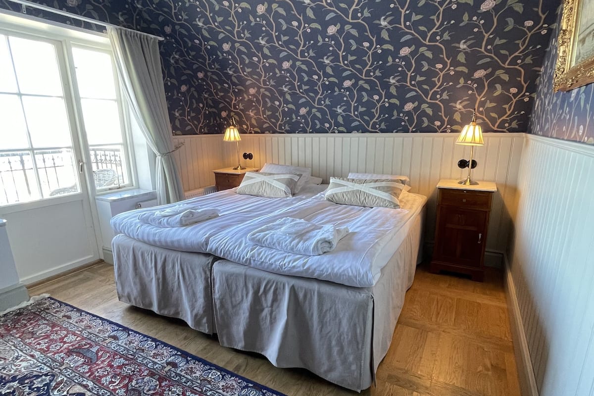 A double bed with white bedding, two bedside tables with lamps, a patterned rug, and a window with curtains.