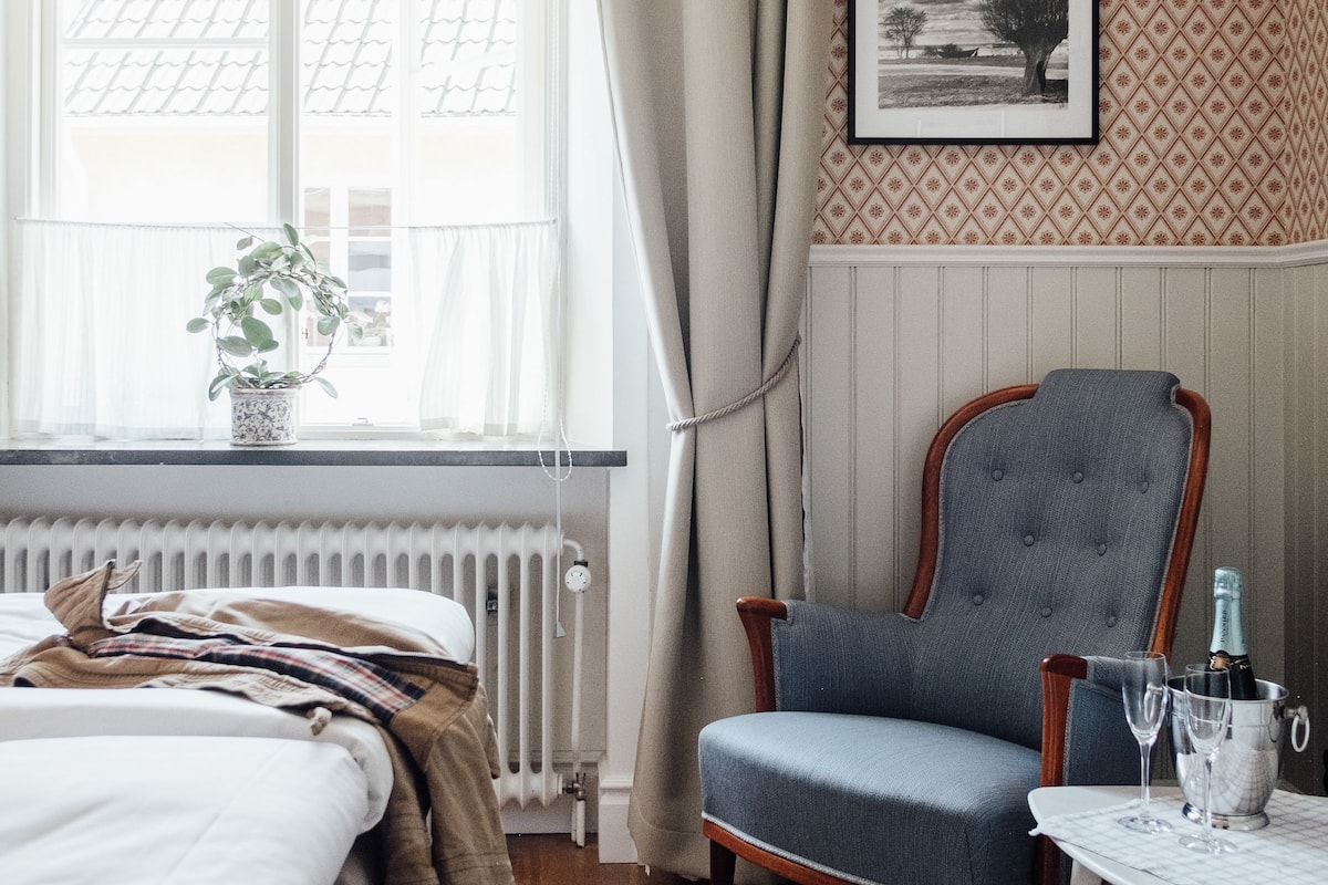 A bedroom with a bed, a chair, a table with glasses, a plant, and a window with curtains.