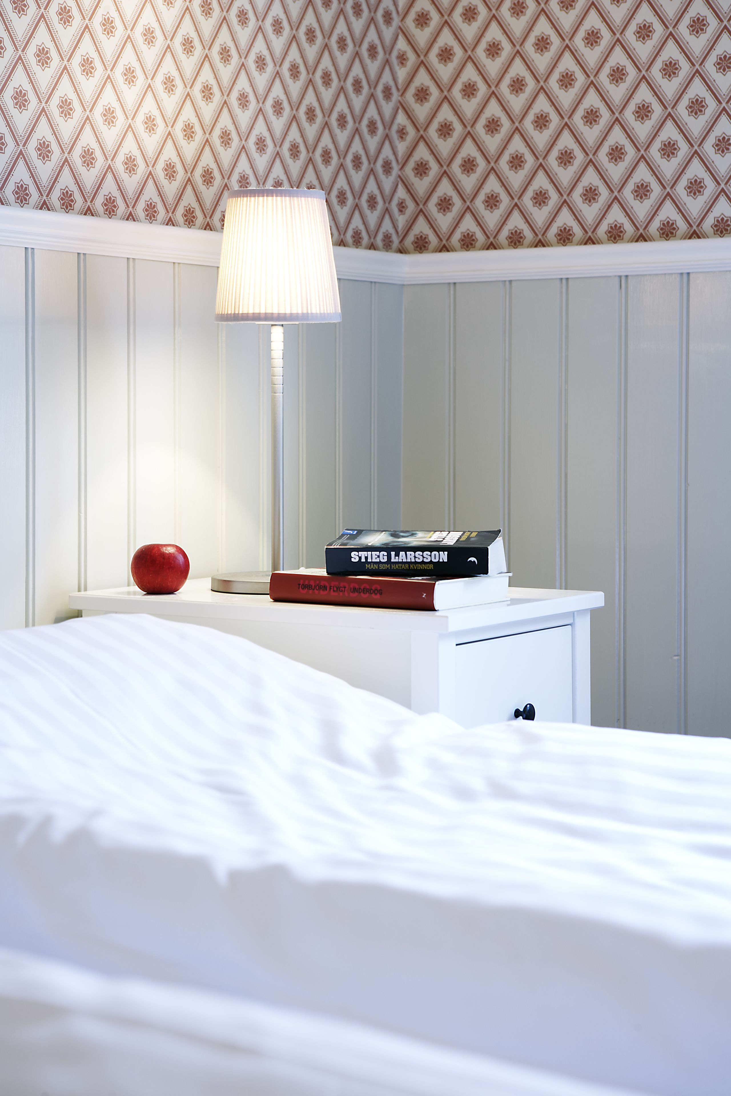 A neatly made bed with a white headboard, a bedside table with a lamp, a book, and an apple.