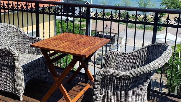 A balcony with wicker furniture and a wooden table, overlooking a body of water. - Kivikstrand Badhotell (Kivik)