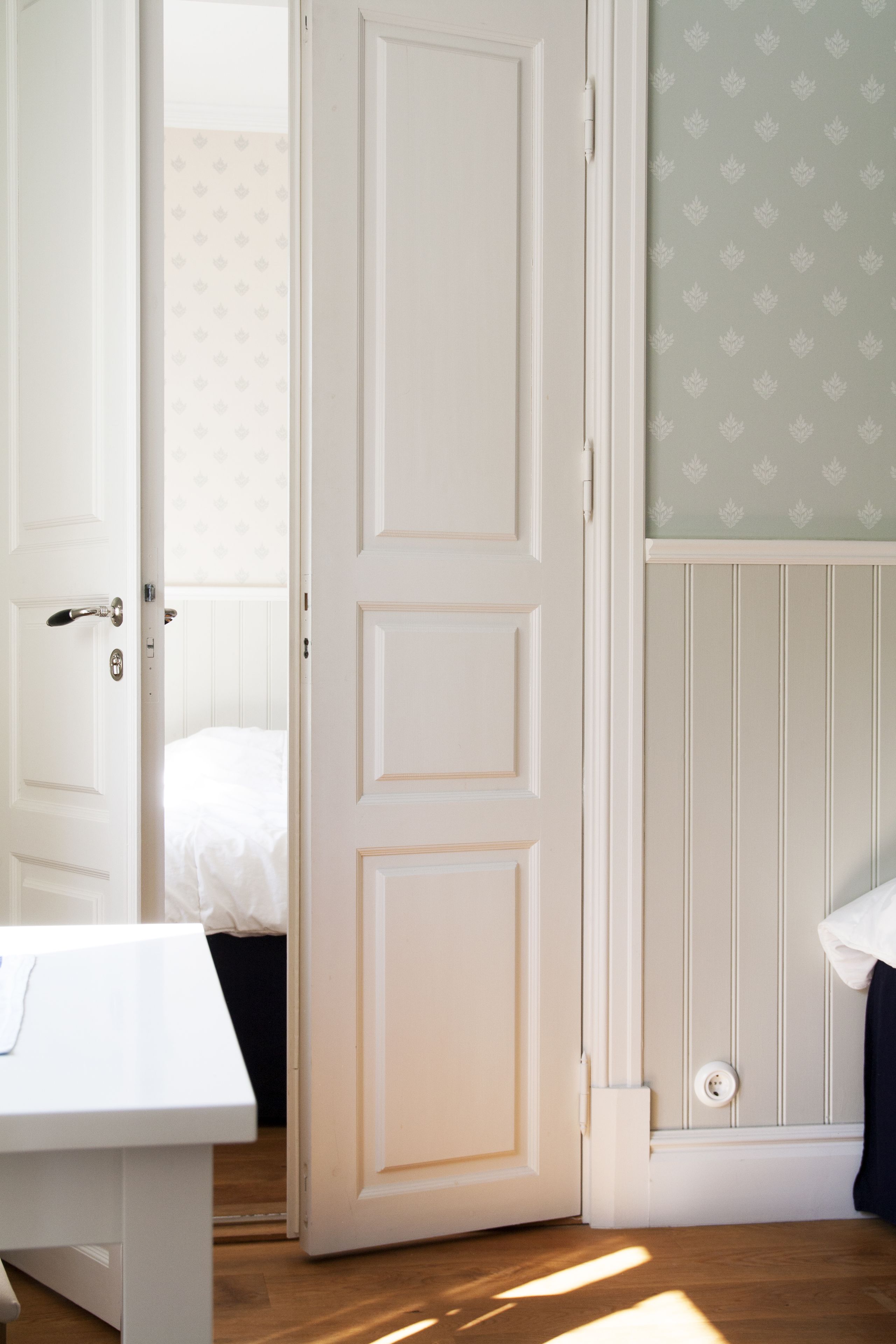 A room with a white door, a bed with white bedding, a white dresser, and a wall with a leaf-patterned wallpaper.