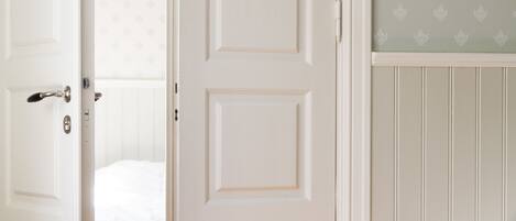 A room with a white door, a bed with white bedding, a white dresser, and a wall with a leaf-patterned wallpaper.
